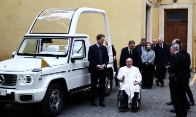 Pope in wheelchair beside popemobile.