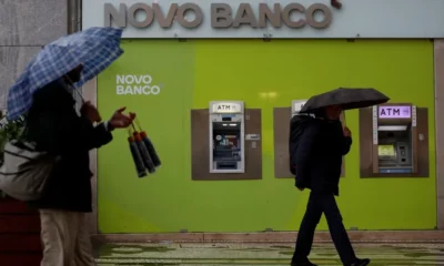 People with umbrellas walking past Novo Banco ATMs.