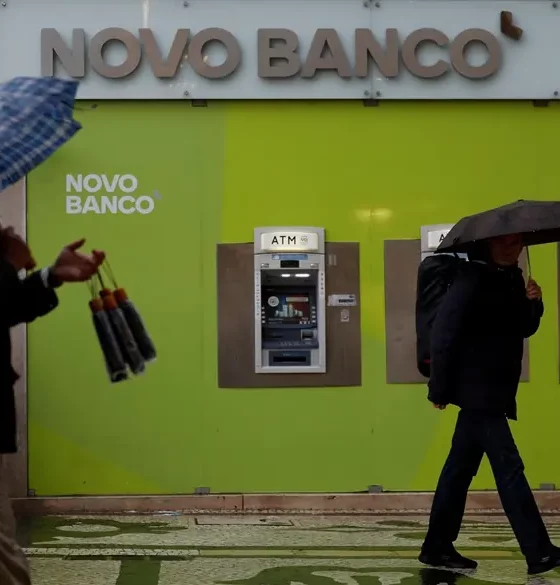 People with umbrellas walking past Novo Banco ATMs.